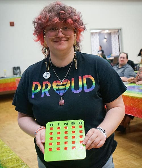 A student holds up a completed bingo card and smiles. They are wearing a rainbow shirt that reads "Proud"