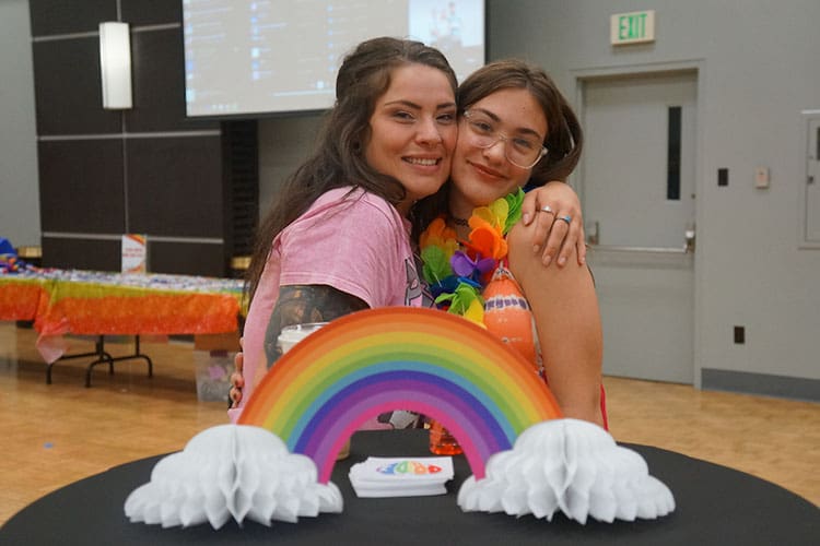 Two students hug. They are standing behind a rainbow display