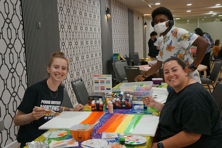 Two people are sitting and painting. One person is standing off to the side wearing a mask