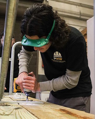 A student wearing long sleeves and protective glasses uses a drill